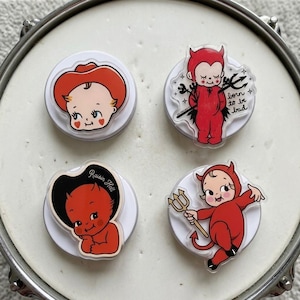 May include: Four circular phone grips featuring illustrated characters. One shows a baby in a red cowboy hat, another a red devil baby with the text "born to be bad", a third a red devil baby with "Raisin Hell", and the fourth a red devil baby holding a trident.