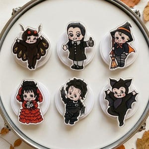 May include: A set of six decorative pins featuring cartoon-style characters: a moth, a girl with braids holding a bottle, a witch with a broom, a girl in a red dress, a character with scissor hands, and a bat, all displayed on white circular bases.