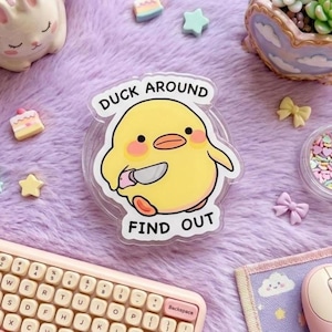 May include: A clear acrylic charm featuring a yellow cartoon duck with pink cheeks holding a small knife. The text DUCK AROUND and FIND OUT is printed in black above and below the duck. The design has a white border and a glossy finish.