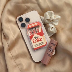 May include: A light-colored smartphone, a clear plastic case containing three small Diet Coke cans labeled Fridge Cigs, a beige fabric scrunchie, and a tube of Trend It Up Pure Nude lipstick with a semi-matte finish are arranged together.