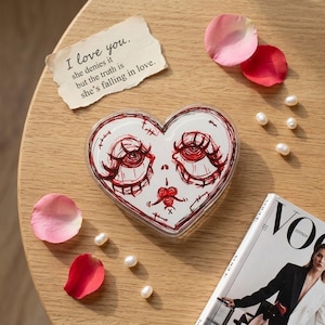 May include: A heart-shaped clear acrylic plaque featuring a red ink sketch of a face with large eyes and long lashes. A torn piece of paper reads: I love you. she denies it but the truth is she's falling in love. Pink rose petals and white pearls.