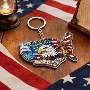 May include: A patriotic keychain in the shape of the United States, featuring a bald eagle, the Liberty Bell, and the American flag. The keychain commemorates the 250th anniversary of the United States in 2026. The keychain is attached to a silver keyring.