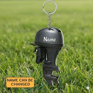 May include: A black outboard motor keychain with a silver keyring and chain. The word "Name" is printed in white on the motor's housing. A yellow rectangle at the bottom reads "NAME CAN BE CHANGED". The keychain is set against a backdrop of green grass.
