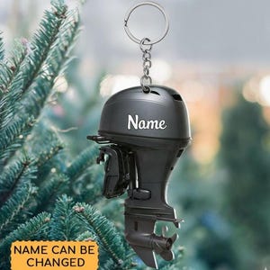 May include: A dark gray outboard motor keychain featuring a silver metal ring and chain. The side of the motor housing displays the white text "Name" in a script font. The design includes mechanical details like the propeller and mounting bracket.
