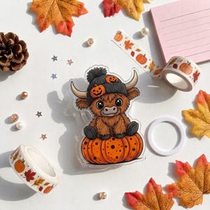 May include: A clear acrylic charm featuring a brown Highland cow wearing a dark gray knit hat with orange jack-o'-lantern accents, sitting atop a large orange pumpkin with black spots, surrounded by autumn leaves, washi tape, and beads.