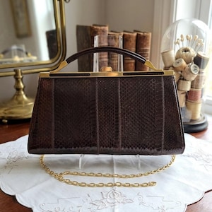 Può includere: Una borsa vintage marrone con un motivo a pelle di serpente. La borsa rettangolare ha una chiusura e attacchi per manici color oro. È visibile una tracolla a catena dorata. La borsa è esposta su un panno bianco ricamato.