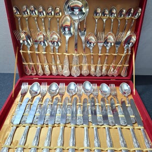 Puede incluir: Un juego completo de cubiertos plateados exhibido en un estuche forrado de terciopelo rojo. El juego incluye cucharas, tenedores, cuchillos y una cuchara de servir. Los mangos tienen un diseño ornamentado y detallado. El estuche está abierto, revelando la disposición organizada de la plata.