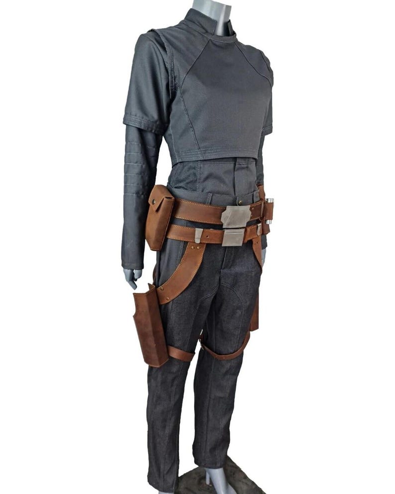 May include: A gray costume with a high collar, short-sleeved top, and long sleeves. The costume includes dark gray pants, a brown leather belt with a silver buckle, and brown leather holsters. The outfit is displayed on a mannequin.