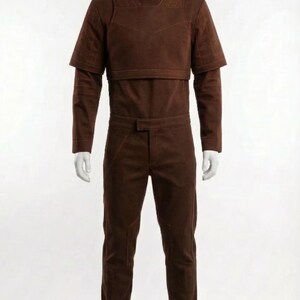 May include: A brown jumpsuit displayed on a mannequin. The outfit includes a high-neck top with shoulder accents, a cropped overlay, long sleeves, and matching pants. Brown boots complete the look. The outfit is a solid color.