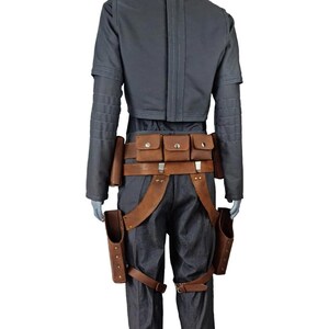May include: A gray costume with a cropped jacket, dark pants, and a brown leather belt with pouches. The costume includes brown leather holsters and straps, creating a rugged, utilitarian look.