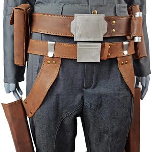 May include: A brown leather belt with a silver buckle and matching pouches. The belt is worn over gray pants and a gray jacket. The belt has silver accents and is secured with rivets. The pouches are rectangular and appear to be made of the same leather.