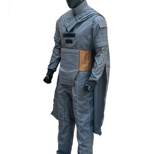 K&ouml;nnte beinhalten: Ein graues Kost&uuml;m mit hohem Kragen, langen &Auml;rmeln und Hose. Das Outfit hat einen braunen G&uuml;rtel und wei&szlig;e Nahtdetails. Die Schaufensterpuppe tr&auml;gt schwarze Handschuhe und Schuhe. Das Kost&uuml;m hat ein kapuzenartiges Design.