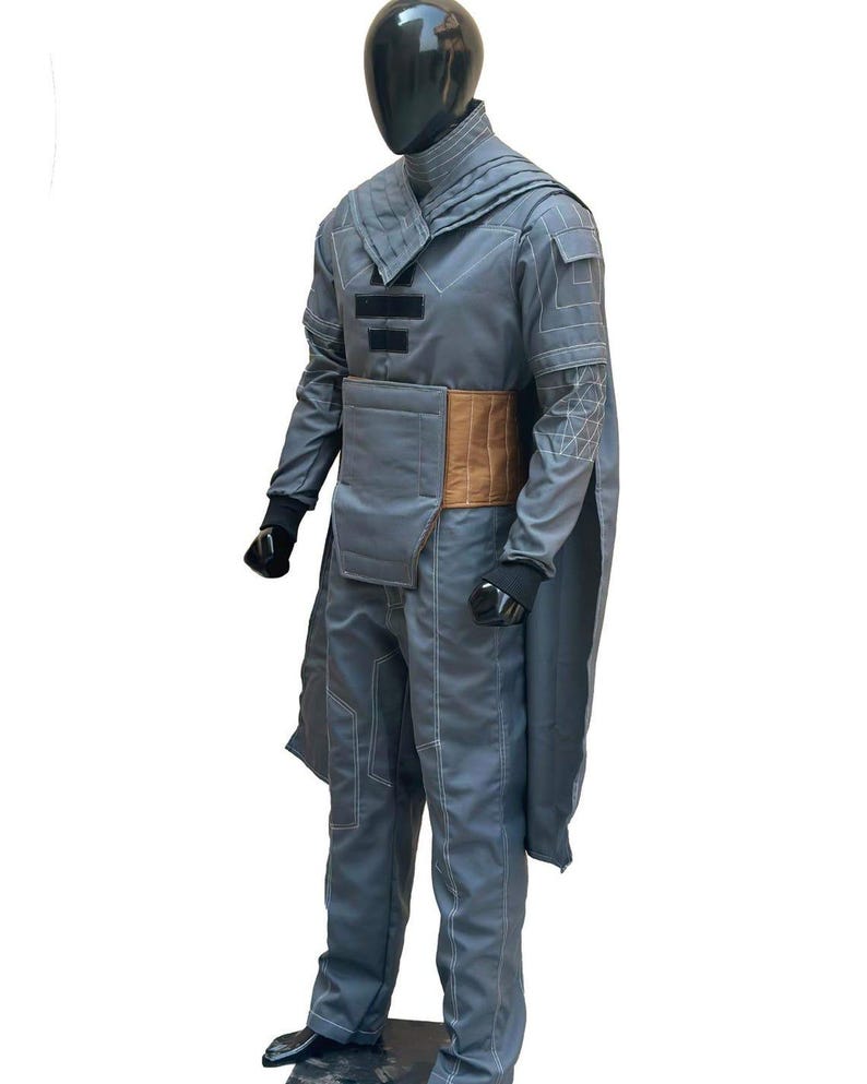 K&ouml;nnte beinhalten: Ein graues Kost&uuml;m mit hohem Kragen, langen &Auml;rmeln und Hose. Das Outfit hat einen braunen G&uuml;rtel und wei&szlig;e Nahtdetails. Die Schaufensterpuppe tr&auml;gt schwarze Handschuhe und Schuhe. Das Kost&uuml;m hat ein kapuzenartiges Design.
