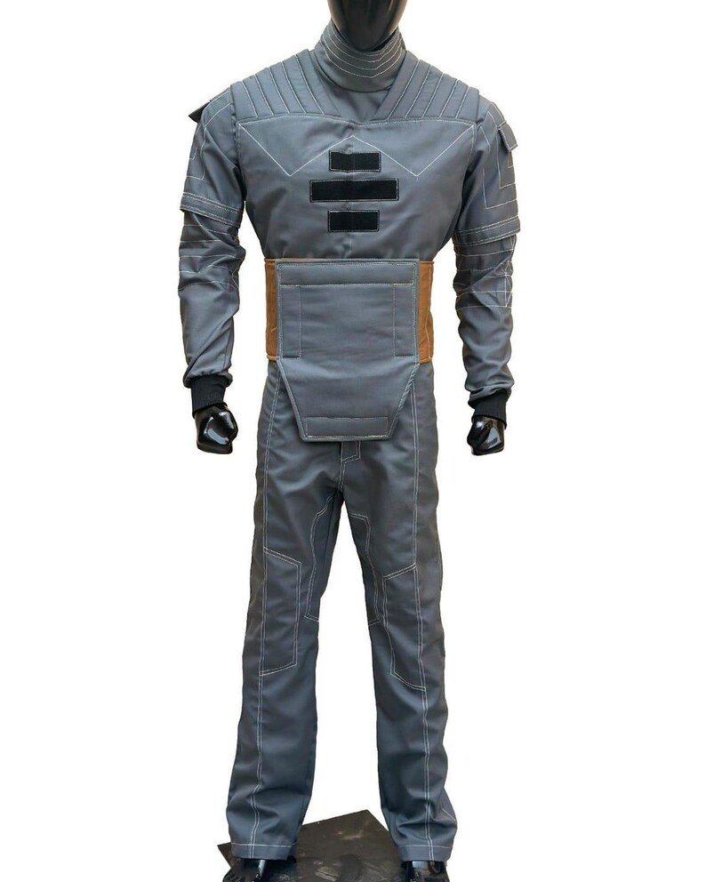 K&ouml;nnte beinhalten: Grauer Overall mit wei&szlig;en N&auml;hten, hohem Kragen und braunem G&uuml;rtel. Das Kost&uuml;m hat gepolsterte Bereiche an Brust und Schultern sowie schwarze Handschuhe. Das Outfit wird auf einer Schaufensterpuppe pr&auml;sentiert.