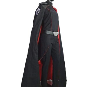 May include: A black costume with a long cape lined in red. The costume includes a black jacket, pants, and belt with a silver buckle. A white emblem is on the left shoulder. The costume is displayed on a mannequin.
