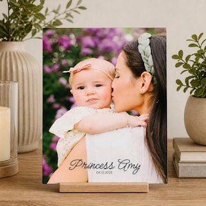 May include: A photo print of a mother kissing a baby, displayed on a wooden stand. The photo features the text "Princess Amy" and the date "04.12.2025". The background shows a blurred floral arrangement.