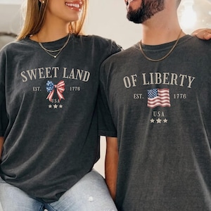 May include: Two dark gray t-shirts with patriotic designs. One shirt says "SWEET LAND" with a bow and stars, the other says "OF LIBERTY" with a flag and stars. Both shirts have "EST. 1776 USA" printed on them.