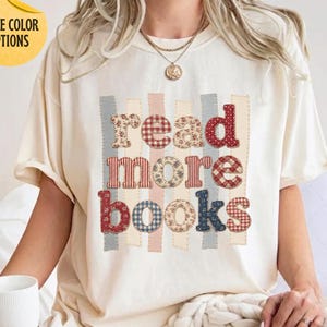 May include: An ivory short-sleeve t-shirt featuring the text "read more books" in a patchwork style with various red, blue, and beige floral and plaid patterns. The letters are arranged over vertical stripes, and the shirt has a crew neckline.