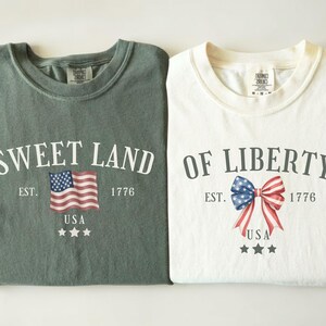 May include: Two folded t-shirts, one green and one white. The green shirt says "Sweet Land" with an American flag graphic. The white shirt says "Of Liberty" with a bow graphic in red, white, and blue. Both shirts have "USA" and stars.