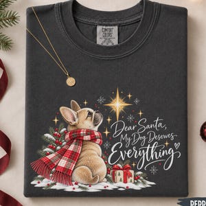May include: A dark gray t-shirt featuring a graphic of a tan French bulldog wearing a red and white plaid scarf. The design includes a bright star, pine branches, a gift box, and the text: Dear Santa, My Dog Deserves Everything.