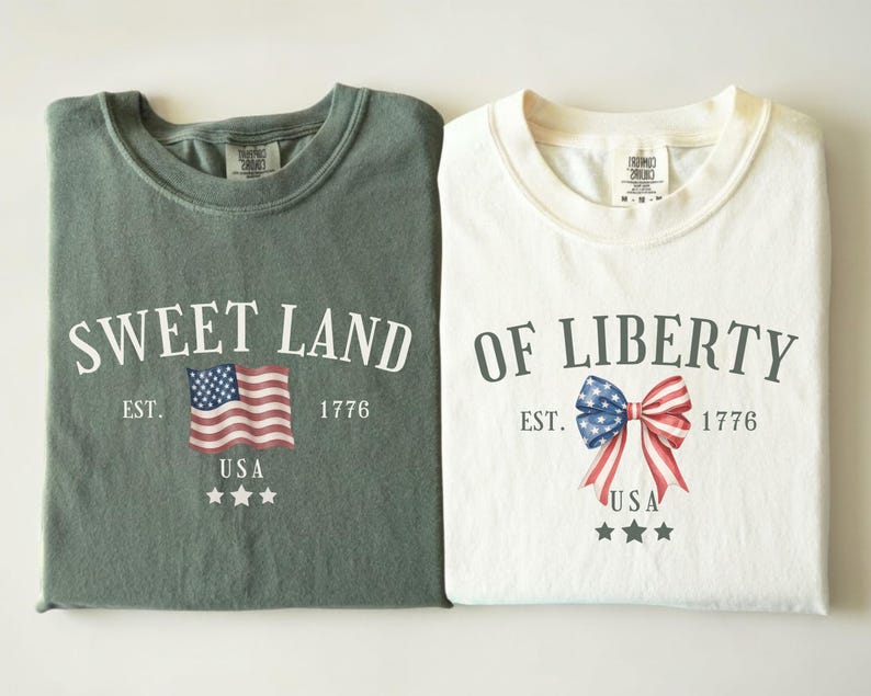 May include: Two folded t-shirts, one green and one white. The green shirt says "Sweet Land" with an American flag graphic. The white shirt says "Of Liberty" with a bow graphic in red, white, and blue. Both shirts have "USA" and stars.
