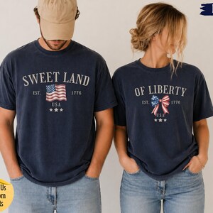 K&ouml;nnte beinhalten: Zwei marineblaue T-Shirts mit wei&szlig;em Text und patriotischen Grafiken. Das linke T-Shirt zeigt SWEET LAND mit einer USA-Flagge, EST. 1776 und drei Sternen. Das rechte T-Shirt zeigt OF LIBERTY mit einer Schleife im USA-Flaggen-Design.