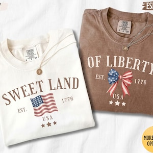 K&ouml;nnte beinhalten: Zwei kurz&auml;rmelige T-Shirts mit Rundhalsausschnitt in Elfenbein und Espresso. Das elfenbeinfarbene Shirt zeigt den Text SWEET LAND, eine US-Flagge, EST. 1776 und drei Sterne. Das Espresso-Shirt zeigt den Text OF LIBERTY, eine Schleife, EST. 1776 und drei Sterne.