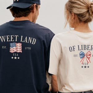 K&ouml;nnte beinhalten: Ein marineblaues T-Shirt mit dem Text SWEET LAND, EST. 1776, USA, drei Sternen und einer US-Flagge sowie ein cremefarbenes T-Shirt mit dem Text OF LIBERTY, EST. 1776, USA, drei Sternen und einer patriotischen Schleife auf der R&uuml;ckseite.