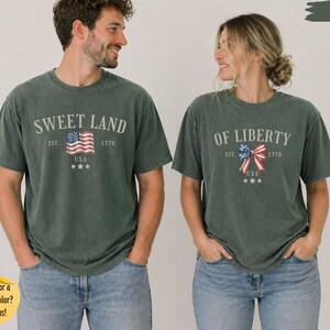 K&ouml;nnte beinhalten: Zwei kurz&auml;rmelige T-Shirts in Moosgr&uuml;n. Das linke T-Shirt zeigt den Text SWEET LAND, EST. 1776, USA und eine Grafik einer amerikanischen Flagge. Das rechte T-Shirt zeigt den Text OF LIBERTY, EST. 1776, USA und eine Grafik einer patriotischen Schleife.