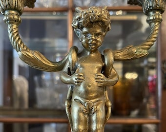May include: A pair of ornate gold-toned metal candle holders with a cherub figure in the center. Each candle holder has a decorative rope-like design and a floral motif at the top.