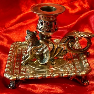 May include: A gold-toned metal candlestick with a winged griffin design. The candlestick has a square base with ornate details and four feet. The candle holder is a simple cup with a decorative rim.