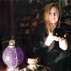 Puede incluir: Una mujer con una camisa negra sostiene una bola de cristal negra en sus manos. Una lámpara de vidrio púrpura con un diseño decorativo se encuentra sobre una mesa frente a ella.