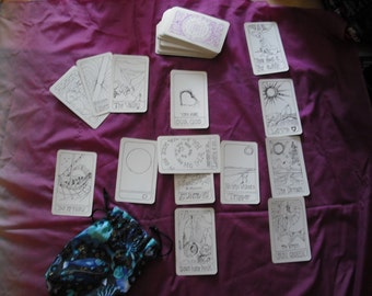 Può includere: Uno spargimento di carte dei tarocchi su una superficie di tessuto viola. Le carte sono bianche con illustrazioni e testo in inchiostro nero. Le carte includono "Il Carro", "La Torre", "Il Mondo", "Il Sole", "La Luna", "La Stella", "L'Eremita", "Il Mago", "L'Imperatrice", "L'Imperatore", "Gli Amanti", "Il Matto", "La Sacerdotessa", "La Ruota della Fortuna", "La Giustizia", "La Temperanza", "La Forza", "La Morte", "Il Diavolo", "L'Appeso", "Il Giudizio" e "Il Mondo".