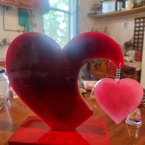 Peut inclure: Une sculpture en forme de cœur rouge avec un petit pendentif en forme de cœur rose suspendu. La sculpture repose sur une base rouge, et le pendentif est attaché par une chaîne argentée. L'arrière-plan est une cuisine.