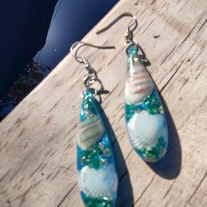 Puede incluir: Un par de pendientes en forma de lágrima con temática playera. Cada pendiente presenta una concha marina encapsulada en resina transparente con detalles azules y verdes. Los pendientes tienen ganchos plateados.