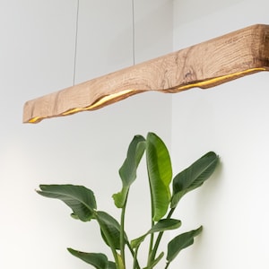 May include: A rectangular wooden pendant light fixture with a natural wood grain finish. The light has a warm, yellow glow emanating from the bottom. The fixture is suspended from the ceiling by thin wires. A green plant is in the foreground.