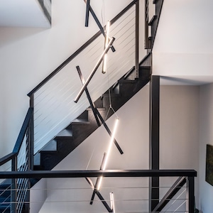 May include: A modern multi-tiered chandelier featuring thin, black linear rods arranged in a crisscross pattern. The fixture includes integrated LED light strips that emit a bright white glow, suspended vertically within a staircase area.