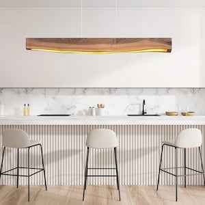 May include: Modern kitchen interior with a wooden light fixture, marble countertop, and three bar stools. The light fixture is a long, rectangular shape with a warm, glowing interior. The bar stools have black metal frames and light-colored seats.