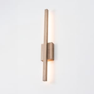 Può includere: Una lampada da parete verticale in legno marrone chiaro con una base rettangolare e una lunga barra centrale sottile. L'apparecchio emette una luce calda e soffusa da dietro la barra di legno, illuminando la superficie della parete.