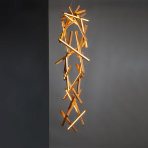 Puede incluir: Lámpara colgante moderna con un conjunto de vigas de madera dispuestas verticalmente. Las vigas emiten una luz cálida y dorada, creando un efecto visual llamativo. La lámpara cuelga de un cable delgado, mostrando un diseño de iluminación contemporáneo.