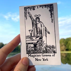 May include: A rectangular white card featuring a black ink illustration of a figure standing at a table with a chalice and pentacle, topped with an infinity symbol. The text at the bottom reads Magician Graves of New York in black font.