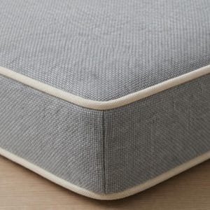 May include: Close-up of a gray cushion with a textured fabric and a cream-colored border. The cushion has a square shape and is placed on a light-colored wooden surface. The fabric appears to be woven.