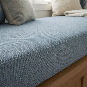 May include: Close-up of a blue upholstered window seat cushion with a textured fabric. The cushion is rectangular and sits atop a wooden base. Soft pillows and a blanket are visible in the background, suggesting a cosy seating area.