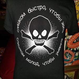 Puede incluir: Camiseta negra con un gráfico blanco de calavera y huesos cruzados. El gráfico tiene el texto "слишком быстро, чтобы жить слишком молод, чтобы умереть" alrededor.