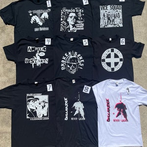 Könnte beinhalten: Neun grafische T-Shirts, hauptsächlich schwarz, mit weißen oder roten Designs. Die Shirts zeigen Bandnamen und Kunstwerke, darunter "The Ruts", "Chaos U.K." und "Discharge". Die T-Shirts liegen auf einer hellgrauen Oberfläche.