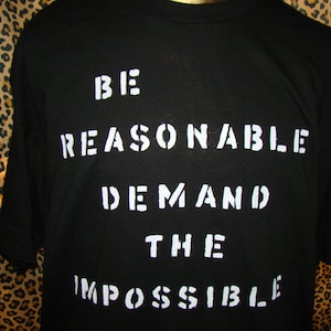 Może przedstawiać: Czarny T-shirt z białym napisem "Be Reasonable Demand The Impossible".