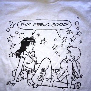 K&ouml;nnte beinhalten: Ein wei&szlig;es T-Shirt mit einer schwarz-wei&szlig;en Illustration von zwei Frauen. Die Frauen sitzen auf dem Boden und eine sagt "This feels good!" in einer Sprechblase. Um die Frauen herum sind Sterne und Kreise.