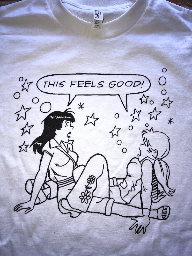 K&ouml;nnte beinhalten: Ein wei&szlig;es T-Shirt mit einer schwarz-wei&szlig;en Illustration von zwei Frauen. Die Frauen sitzen auf dem Boden und eine sagt "This feels good!" in einer Sprechblase. Um die Frauen herum sind Sterne und Kreise.