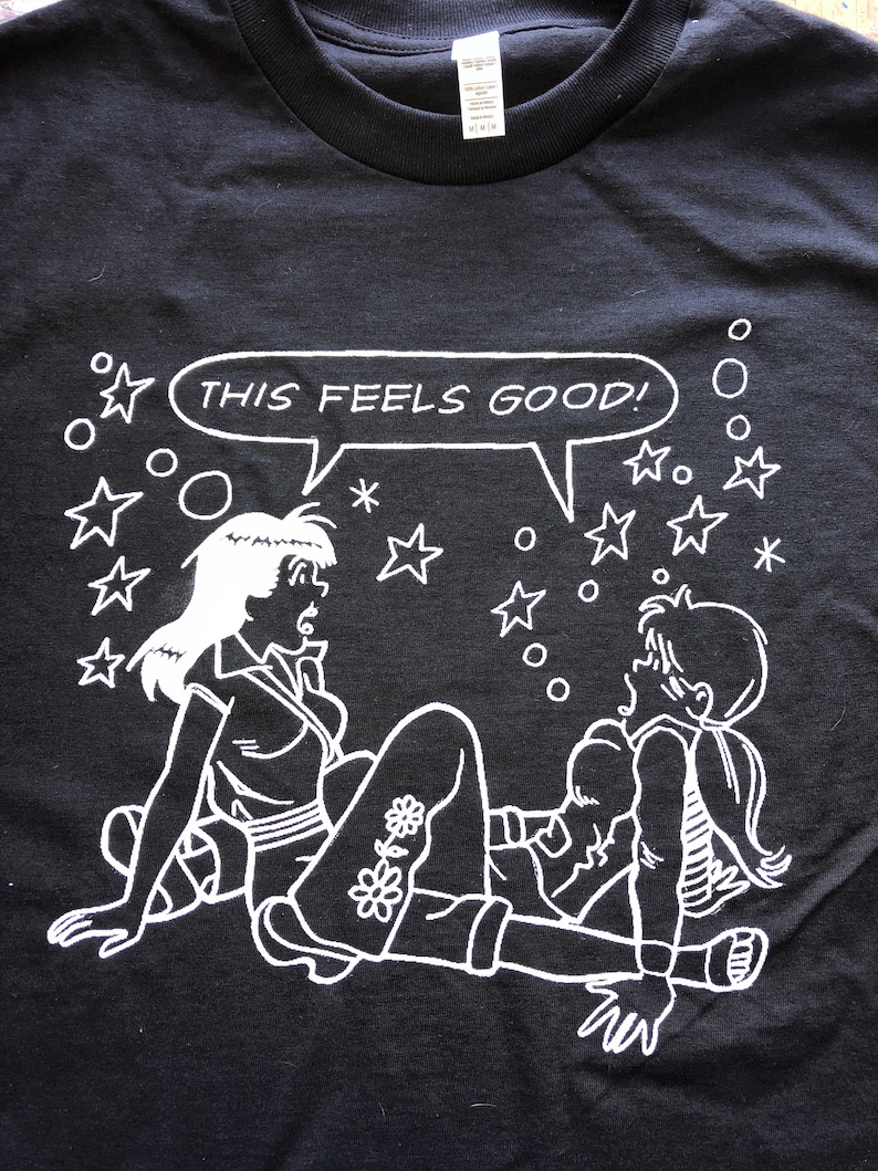 K&ouml;nnte beinhalten: Schwarzes T-Shirt mit einer wei&szlig;en Grafik von zwei Comicfiguren mit dem Text "THIS FEELS GOOD!" in einer Sprechblase. Der Hintergrund ist mit wei&szlig;en Sternen und Kreisen gef&uuml;llt.
