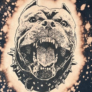 以下が含まれることがあります： 漂白剤のしぶきがかかったデザインの黒いTシャツ。黒いスタッズのついた首輪をした、うなり声をあげているピットブルの白黒の画像が描かれている。